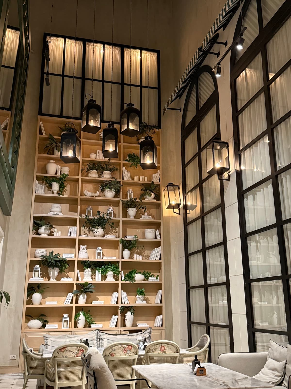 A floor-to-ceiling pale-wood shelving unit at Willow's District 5, stacked with ceramic vases, books and greenery, with black lantern pendants hanging in front of it.