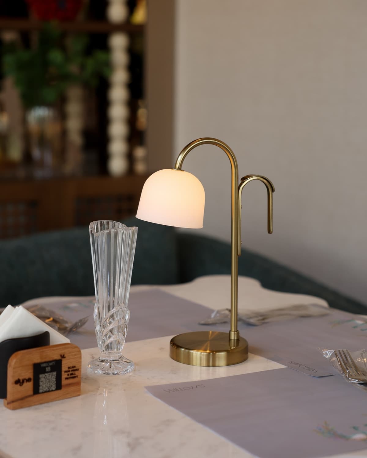 A laid marble table at Willow's Golf Central, with a brass arc lamp, a fluted bud vase and wrapped cutlery on a branded placemat, against a teal banquette.